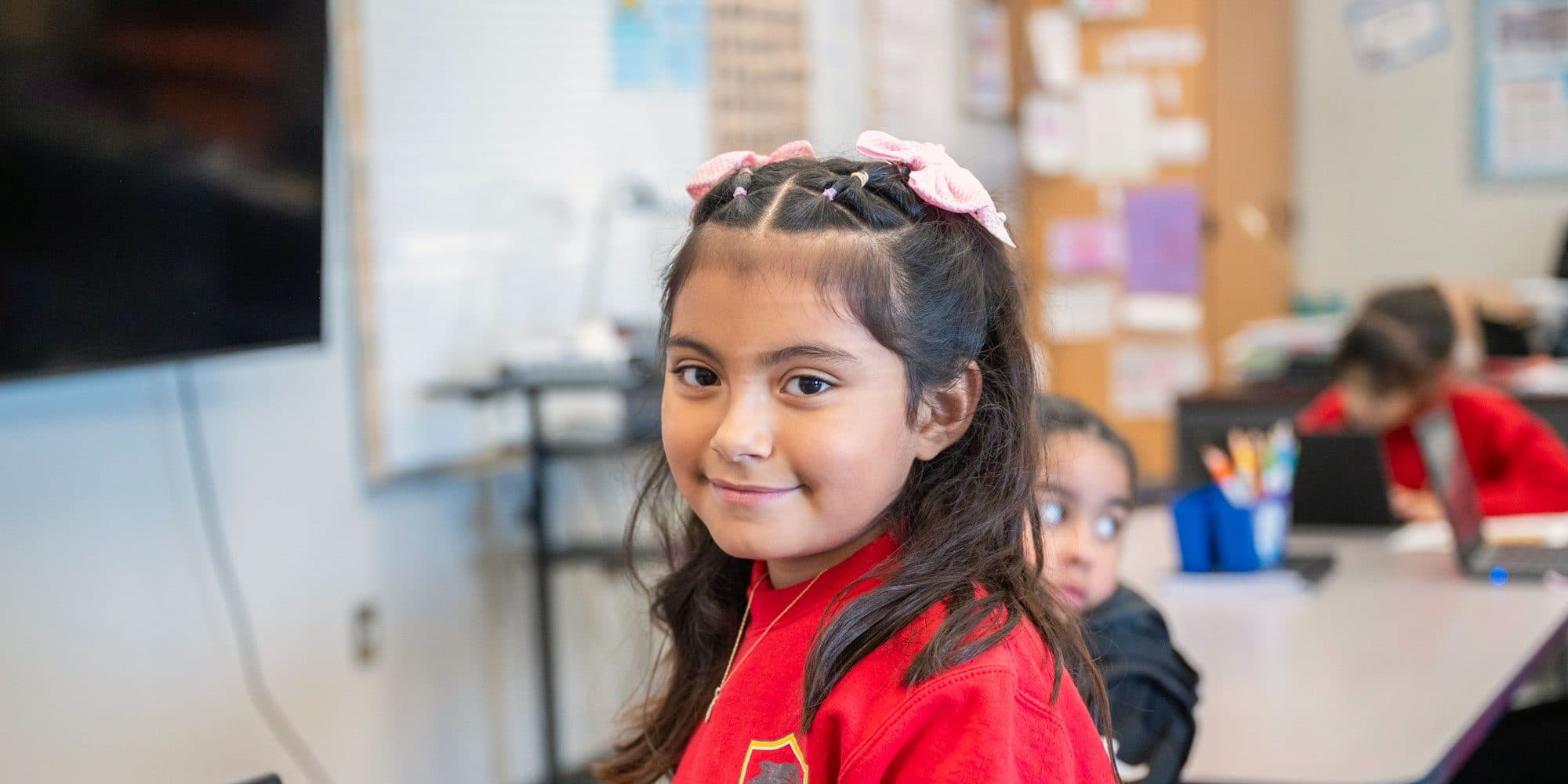 Smiling student with pink bows in a classroom.