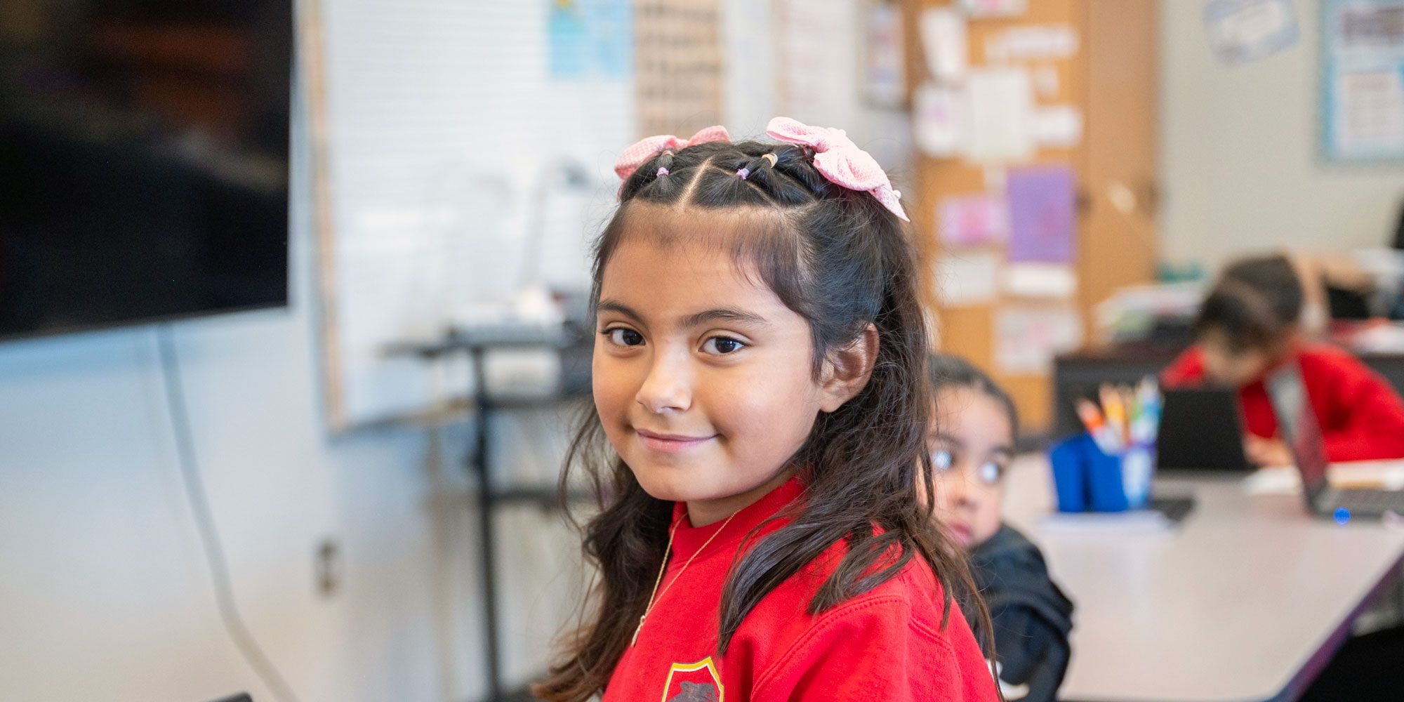 Smiling student with pink bows in a classroom.