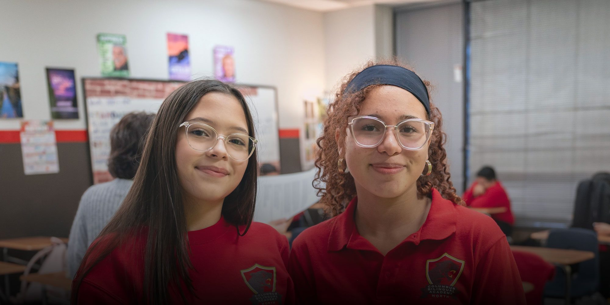 Two smiling students in red Columbus Bilingual Academy North uniforms in a classroom.